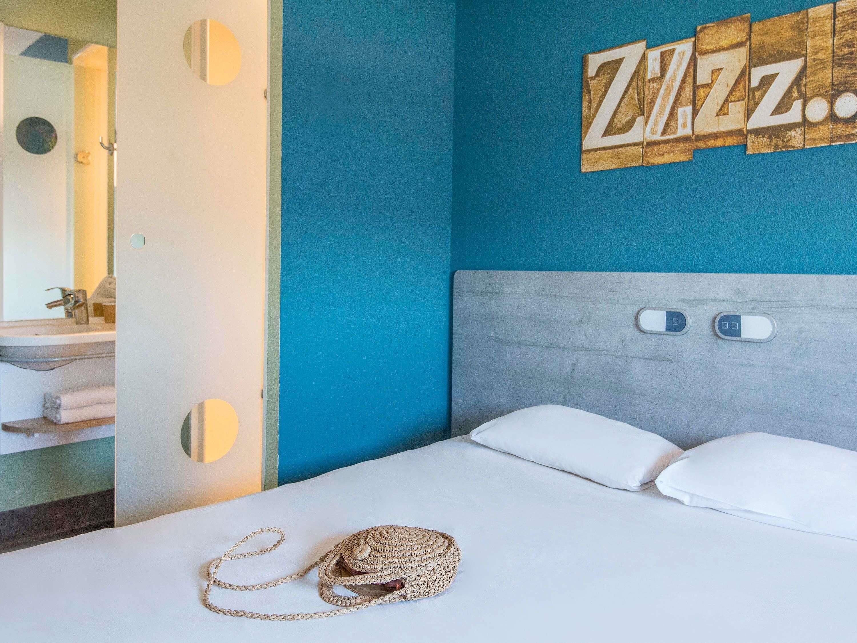 Une chambre à coucher avec un lit, des oreillers, une tête de lit, une table de chevet, une lampe et un mur orné d'un panneau indiquant « Zzz ».
