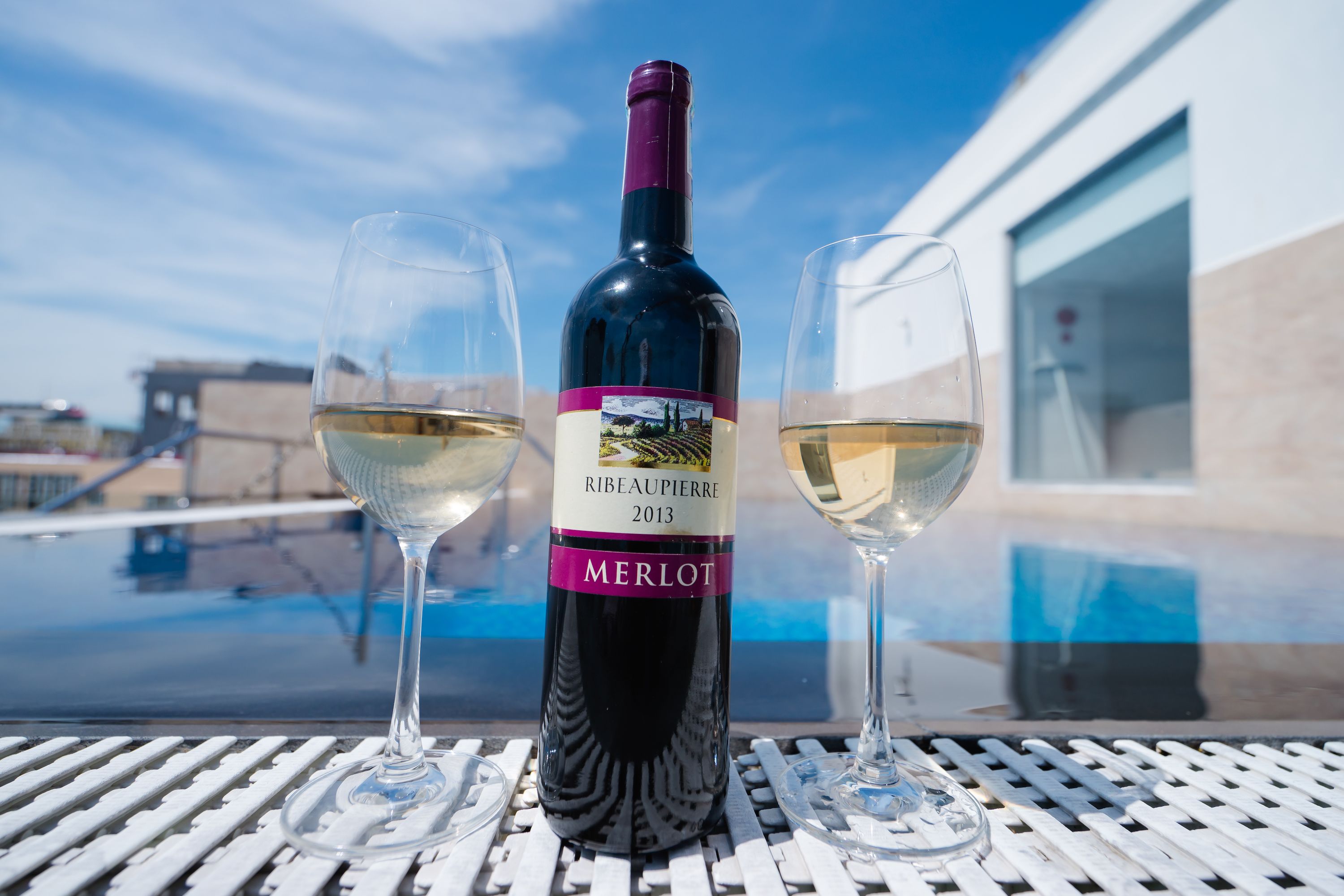 A bottle of 2013 Riebeaupiere Merlot wine and two glasses on a table with a pool in the background.