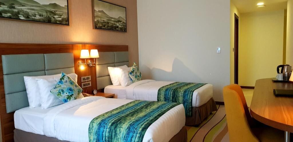 Habitación de hotel con dos camas, cabecero de madera, mesita de noche con lámpara y mesa de comedor con sillas.
