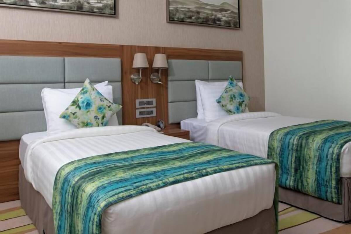 A hotel room with two beds, each with a headboard, white bedding, and a green and blue striped blanket. There are two wall-mounted lamps and a painting above the beds.