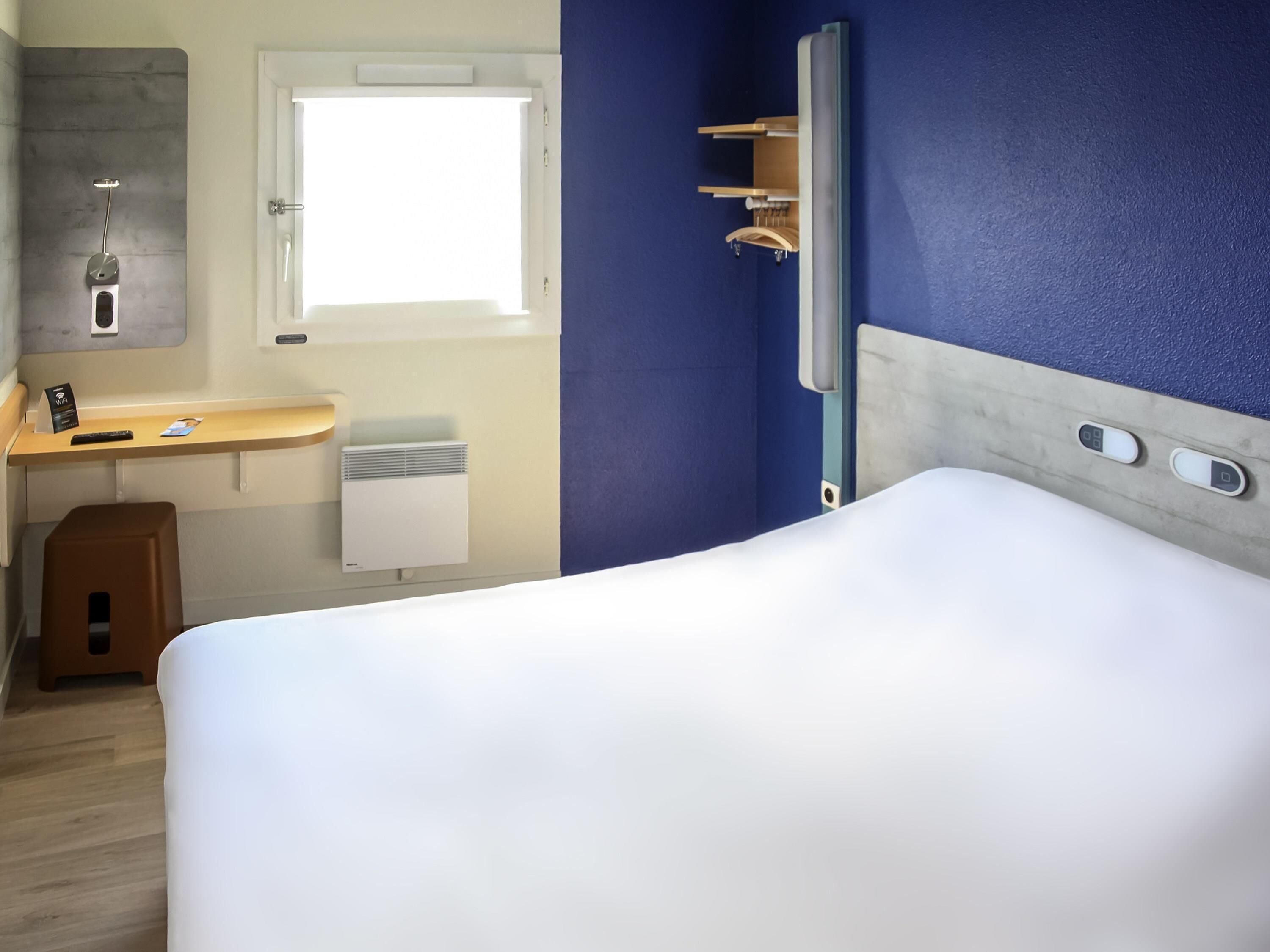 Habitación de hotel con cama, escritorio con silla, ventana, ducha y panel de control.