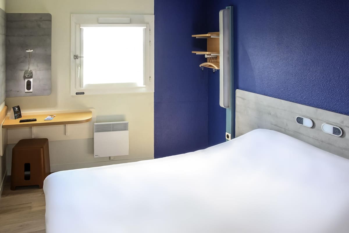 Habitación de hotel con cama, escritorio con silla, ventana, ducha y panel de control.