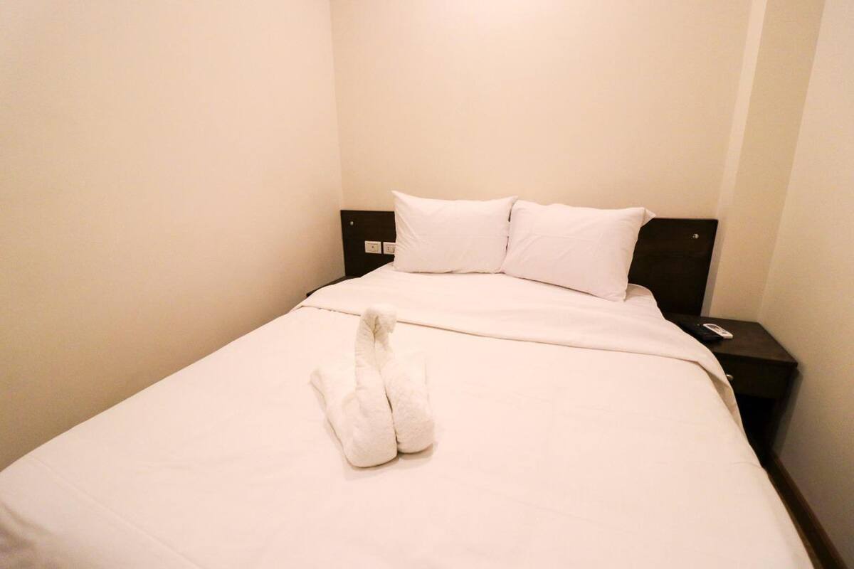 Habitación de hotel con una cama bien hecha, una mesita de noche con un control remoto y un interruptor de luz en la pared.