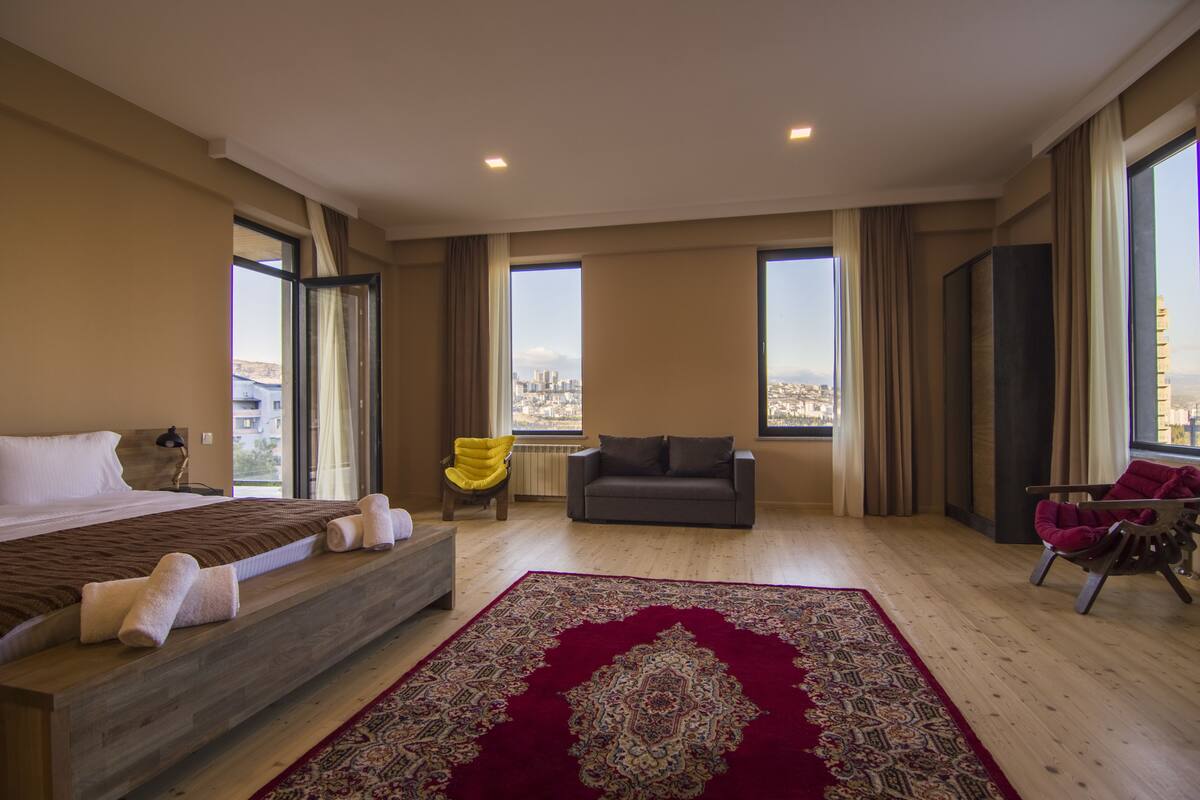 Une chambre d’hôtel spacieuse, dotée d’un grand lit, d’un canapé, d’un tapis à motifs rouges et offrant une vue sur la ville grâce à de grandes fenêtres.