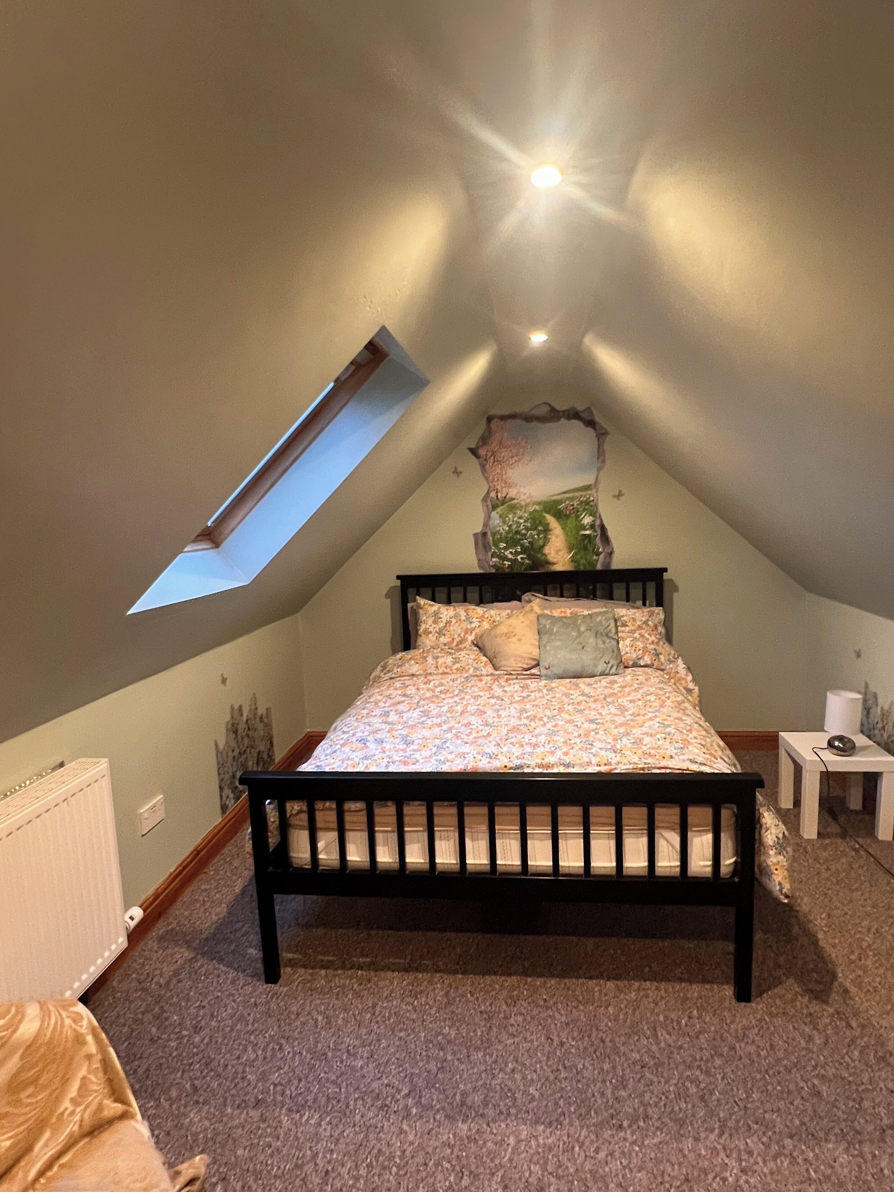 A bedroom with a sloped ceiling, a skylight, a bed with floral bedding, a small bedside table, and a radiator.