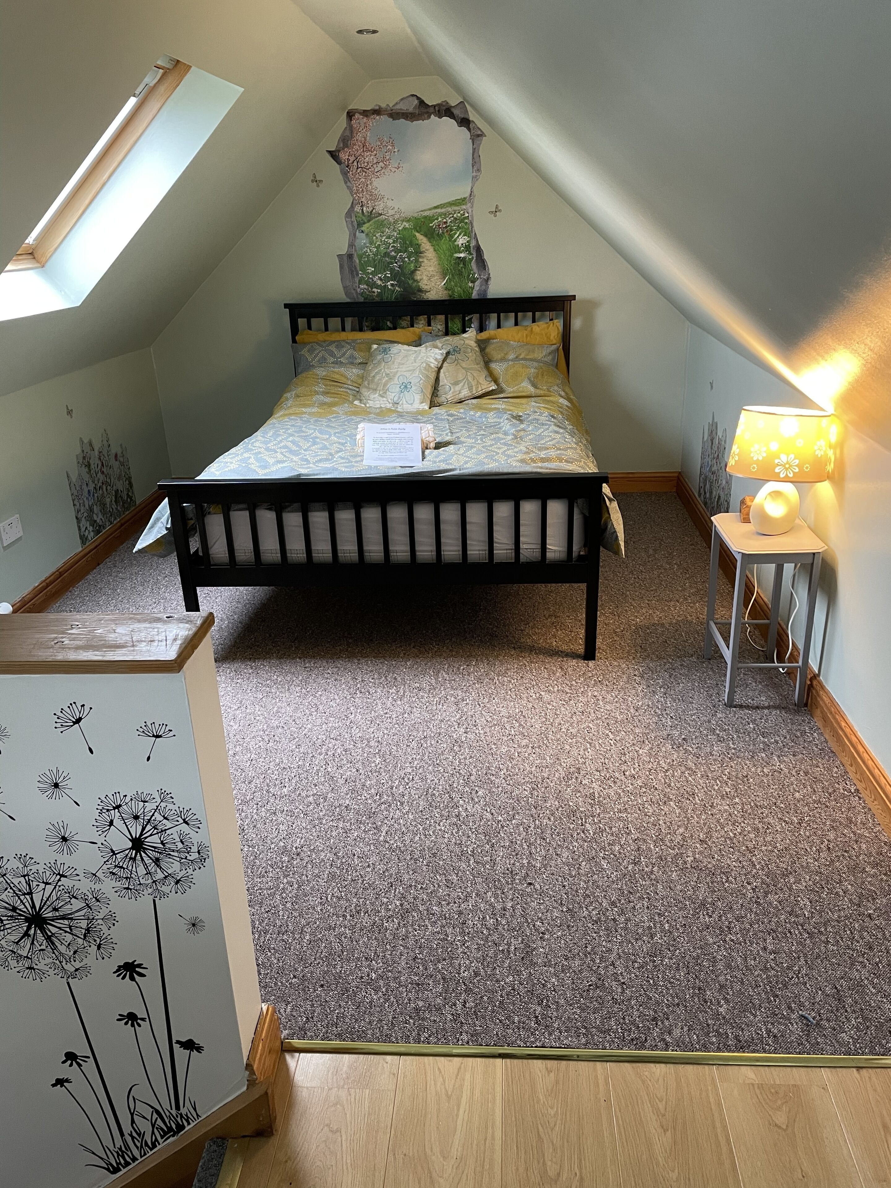 Um quarto com teto inclinado, uma cama com colcha florida, uma mesa de cabeceira com abajur e um mural de parede com um caminho de jardim.