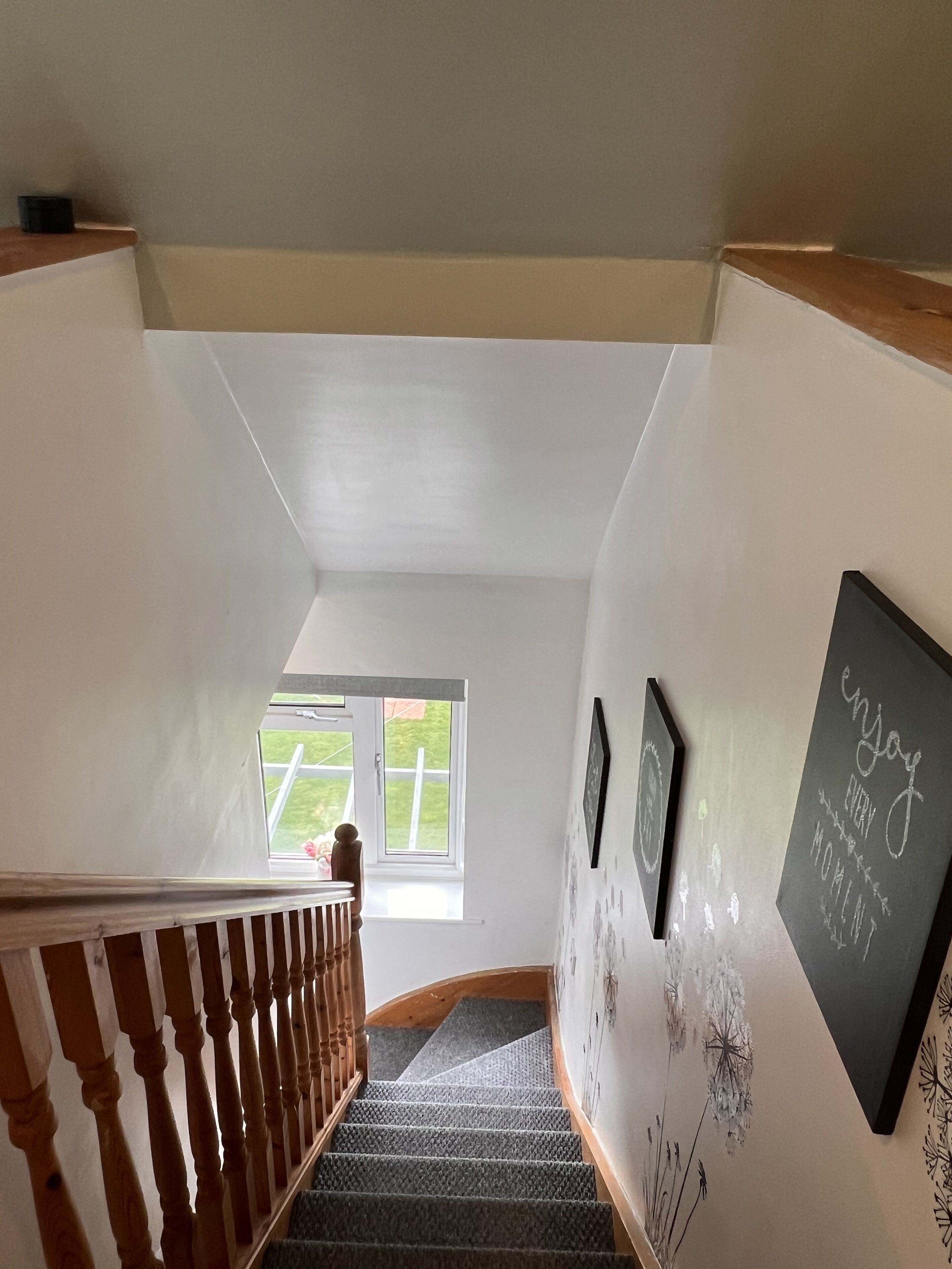 A staircase with a wooden railing, a window with a view of greenery, and a wall with framed pictures and a chalkboard with text.