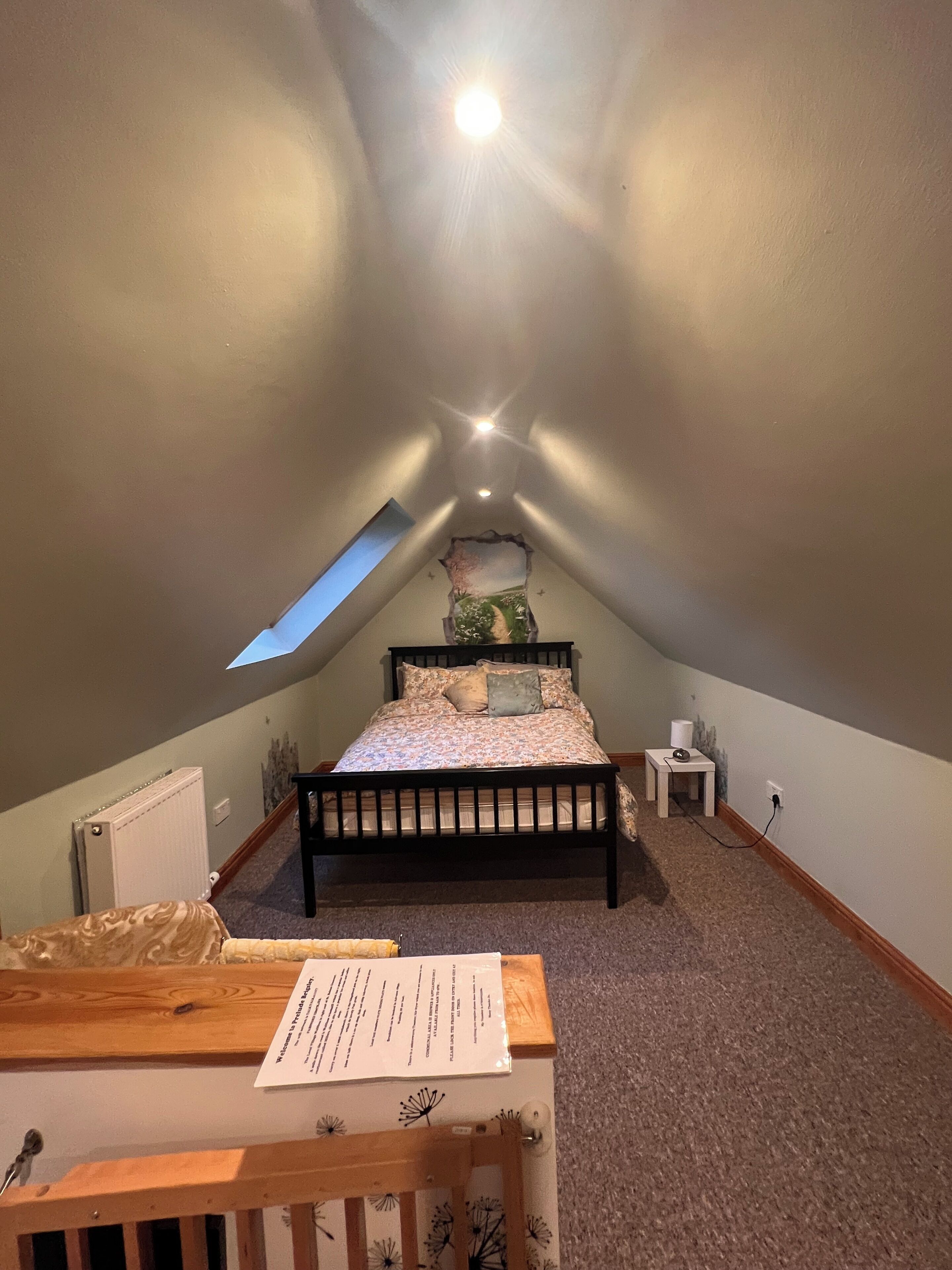 Quarto com teto inclinado, cama, mesinha, radiador e janela.