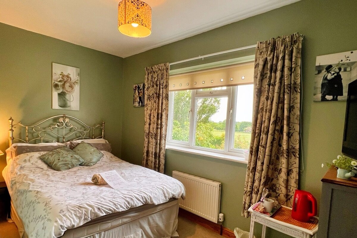 A bedroom with a bed, a window with curtains, a radiator, a bedside table with a kettle, and a picture on the wall.