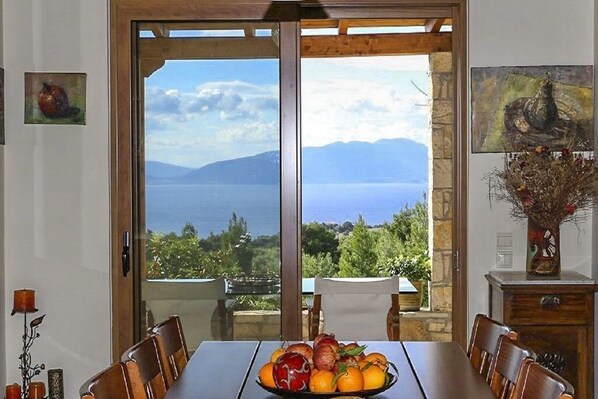 Un coin repas avec une table dressée pour quatre personnes, un saladier rempli de fruits et une vue sur la mer et les montagnes, visible à travers une porte vitrée.