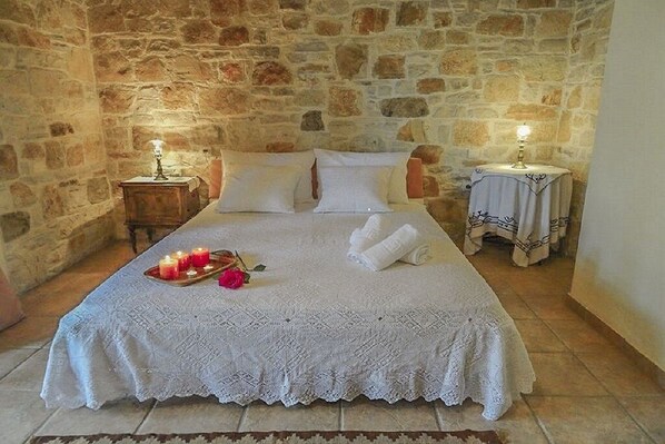 A cozy room with a stone wall, a neatly made bed with a white bedspread, two bedside tables with lamps, and a tray with candles and a rose.
