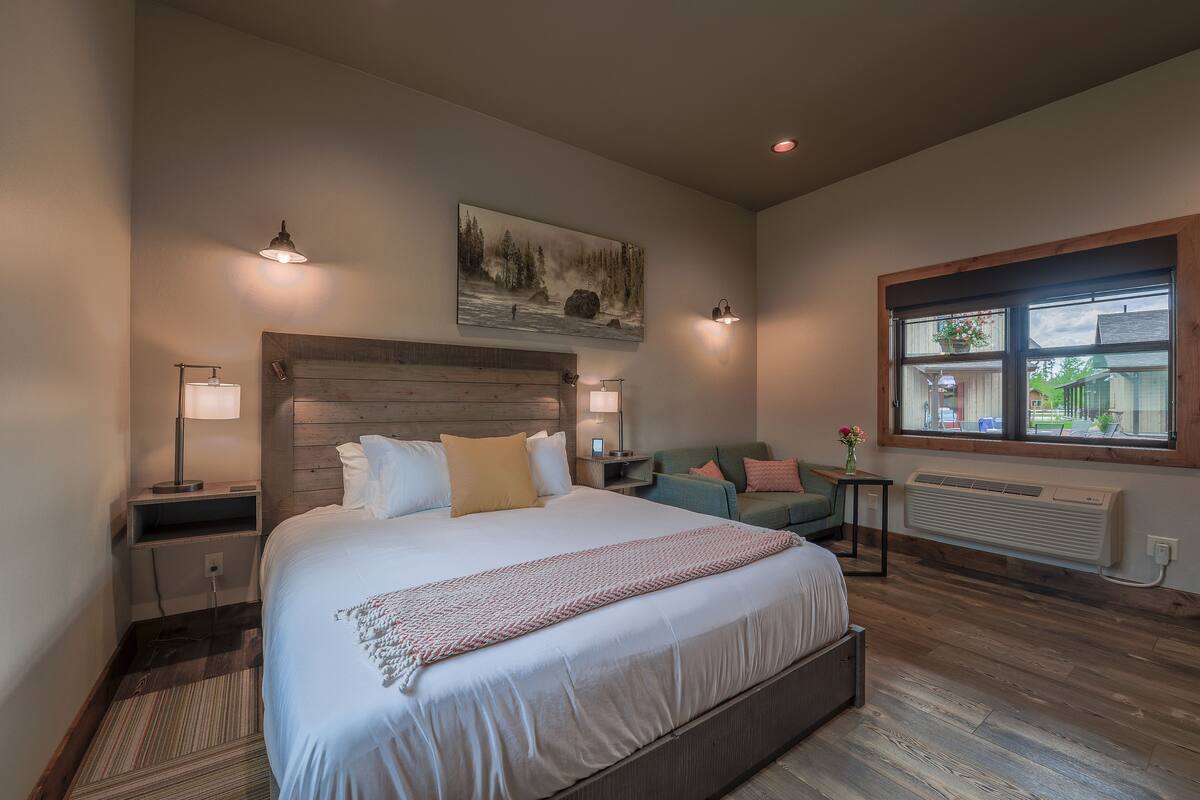 A bedroom with a large bed, a wooden headboard, a nightstand with a lamp, a small table with a vase, and a window with a view of the outside.