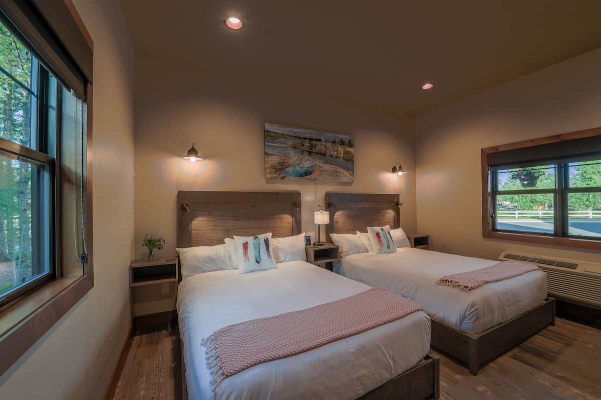 A hotel room with two beds, a painting above the beds, and a window with a view of greenery.