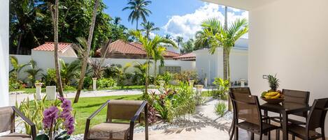 A patio area with outdoor furniture, a ceiling fan, and a view of a garden with palm trees and a building with a red roof.