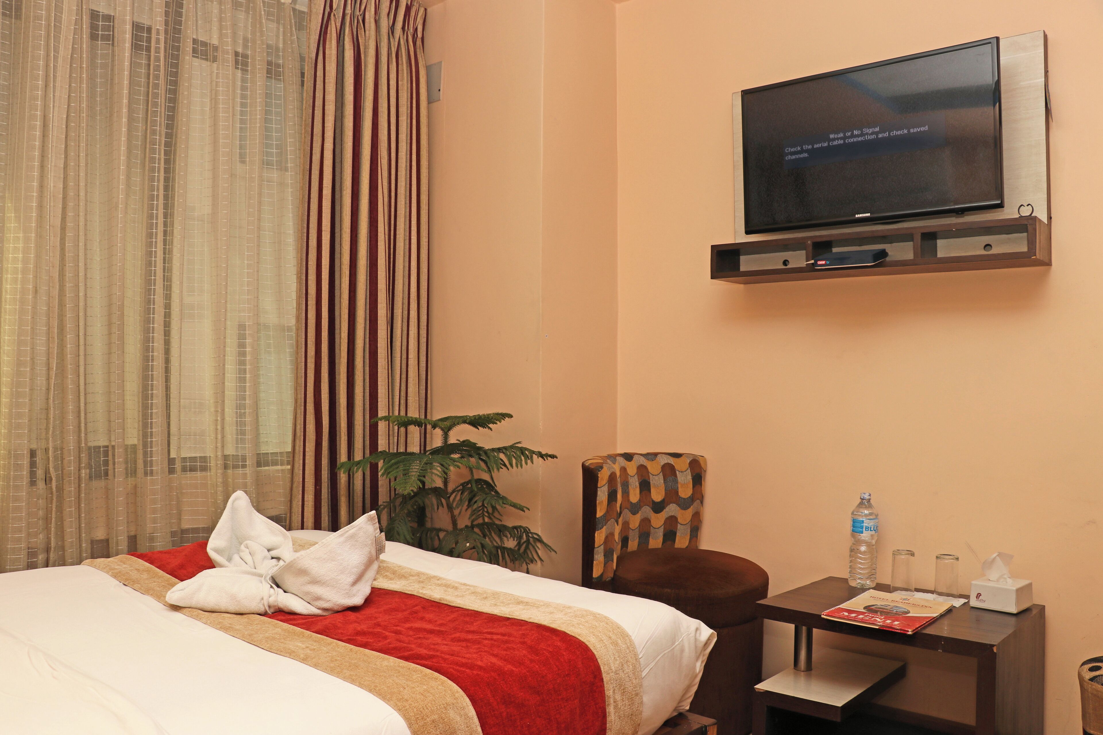 A hotel room with a bed, a TV mounted on the wall, a small table with a bottle and glasses, and a chair.