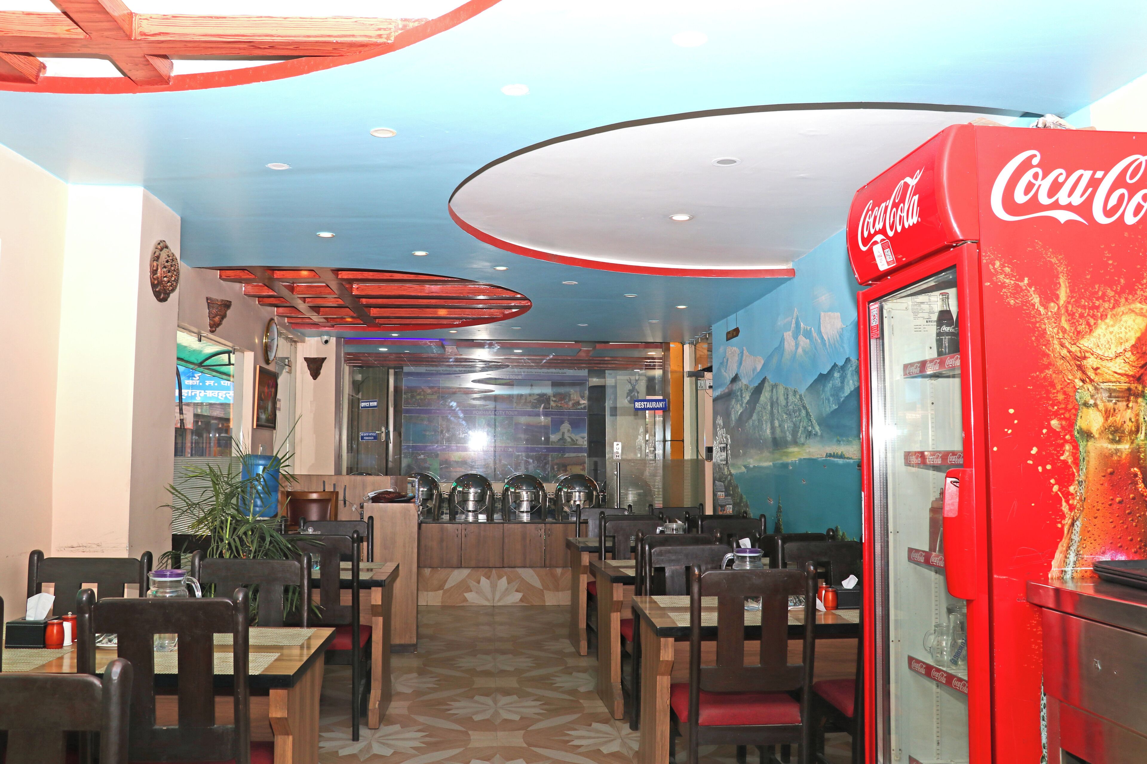 L’intérieur d’un restaurant avec des tables et des chaises en bois, un distributeur automatique de Coca-Cola et une fresque murale.