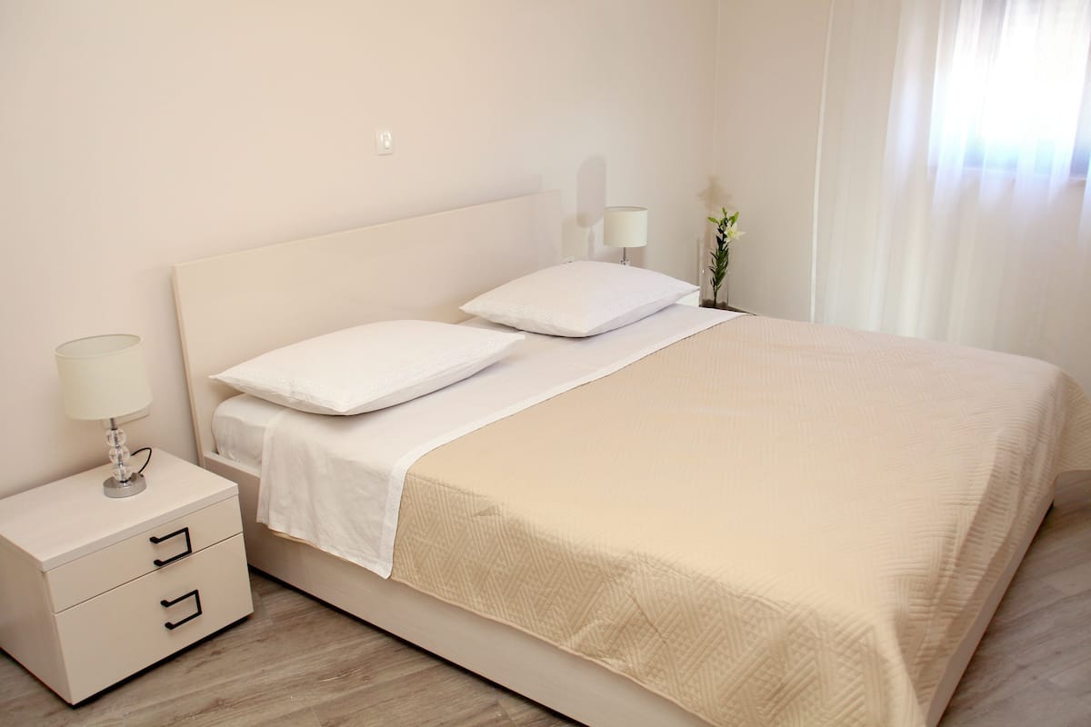 A neatly made bed with a white headboard, a bedside table with a lamp, and a small potted plant.