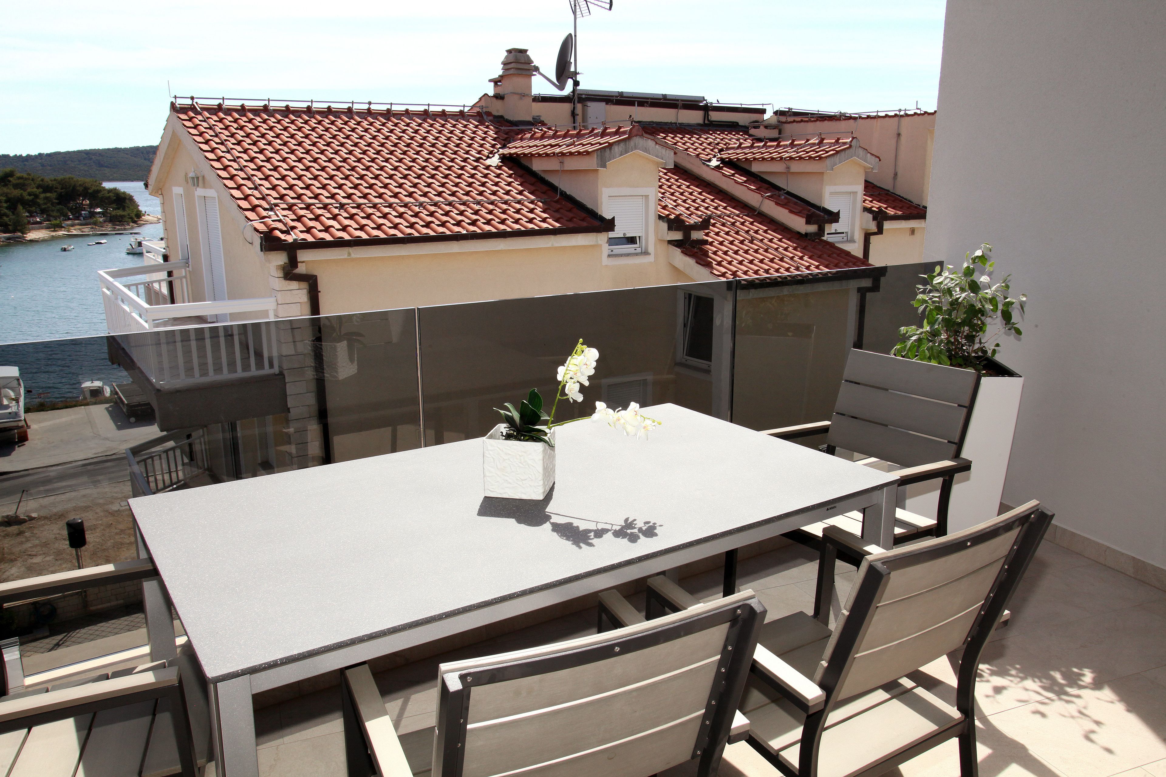 A balcony with a table and chairs, overlooking a coastal area with terracotta-roofed buildings and a body of water.