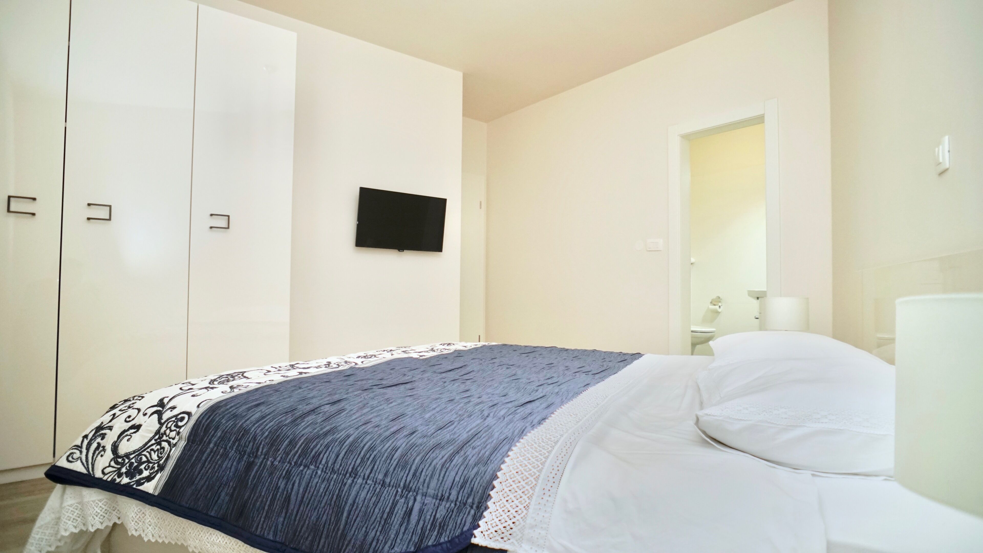A bedroom with a bed, a wardrobe, a flat-screen TV, and a bathroom visible through an open door.