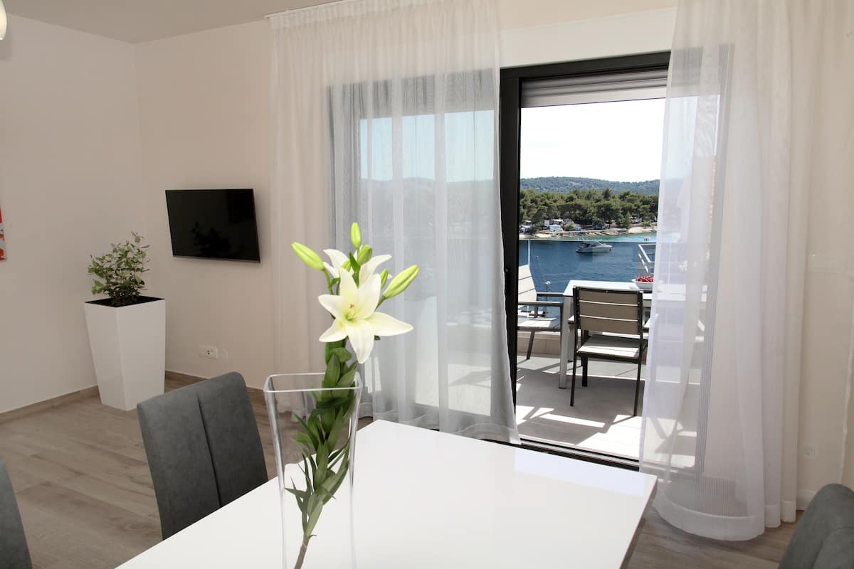 A modern living room with a dining table, chairs, a TV mounted on the wall, and a balcony with a view of the sea.