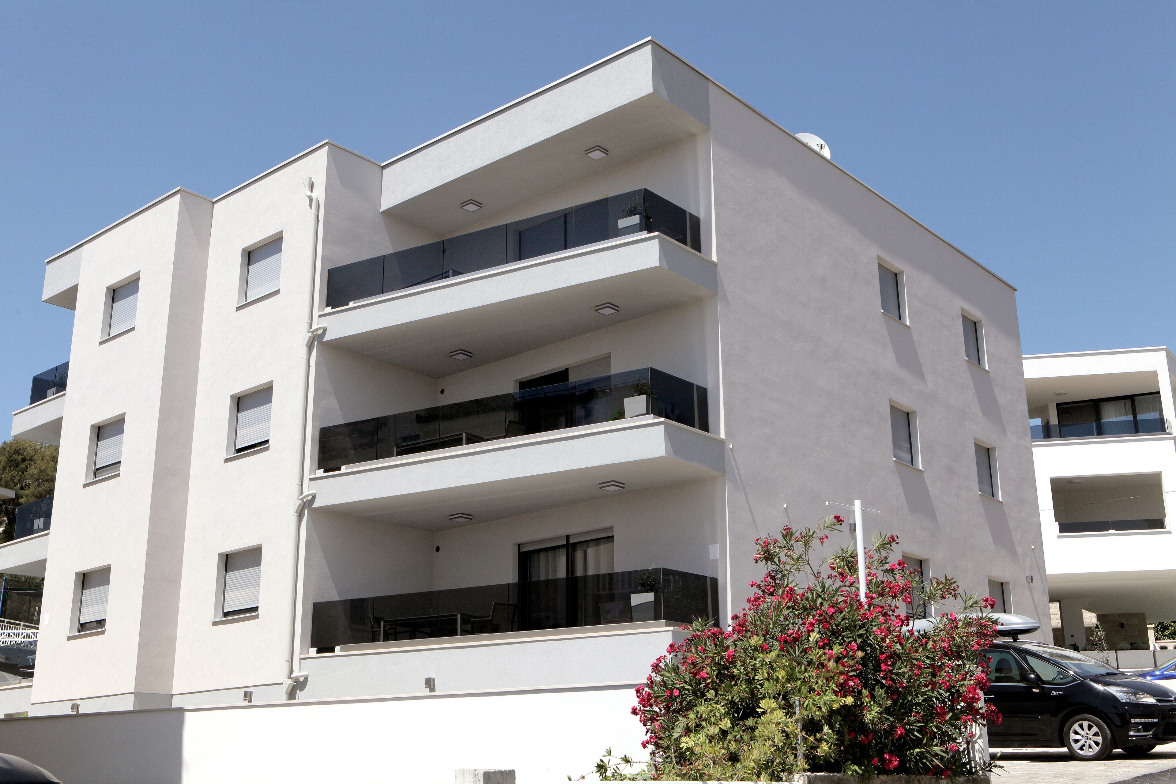 A modern multi-story building with balconies, a car parked in front, and a flower bush in the foreground.