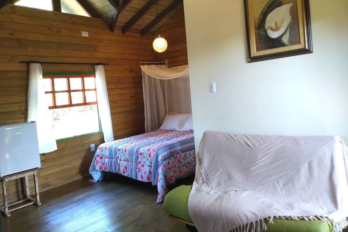 A room with wooden walls, a bed with a floral bedspread, a green chair, a white desk, and a window with curtains.