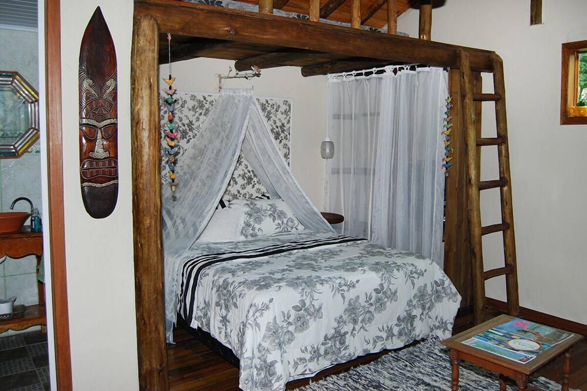 A bedroom with a wooden ceiling, a bed with a floral bedspread, a wooden ladder, and a small table with a magazine.