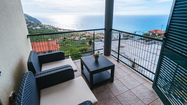 A balcony with wicker furniture, a small table with a vase, and a view of the sea and distant buildings. - Sea View Apartment Single Beds with Swimming Pool (Arco da Calheta)