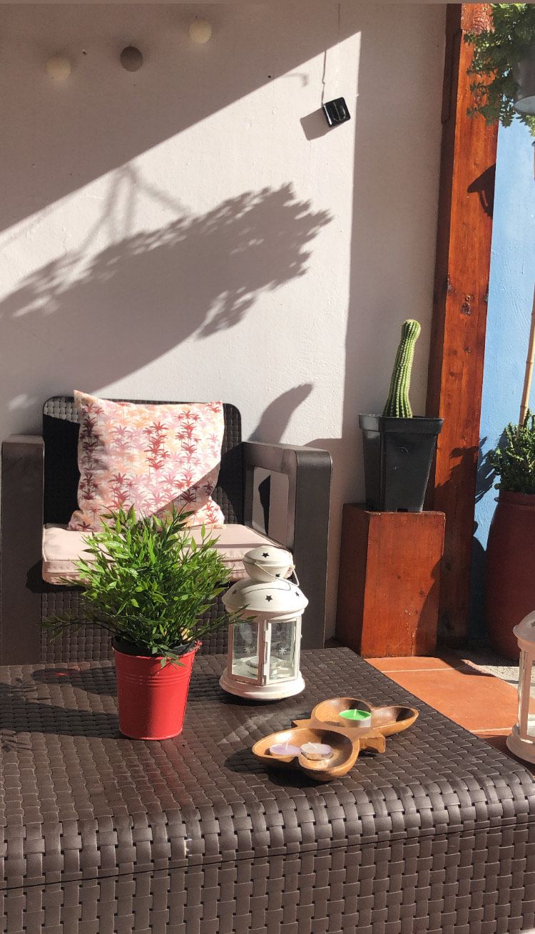 Ein sonniger Außenbereich mit Sitzgelegenheit, einem gewobenen Tisch, einem Topfpflanzen, einem weißen Laternen und einer Holzschüssel.