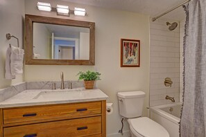 Un bany amb un lavabo de marbre, un tocador de fusta, un vàter, una dutxa amb porta de vidre i un quadre al mur.