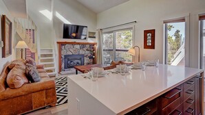 Une cuisine moderne avec une cheminée en pierre, un coin repas avec une table dressée pour quatre personnes et une grande fenêtre donnant sur la forêt.