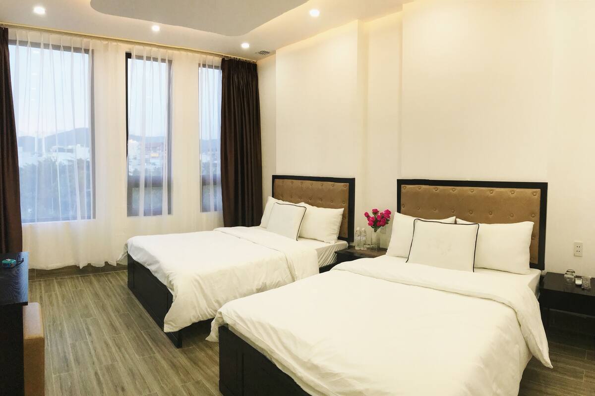 Habitación de hotel con dos camas, una mesita de noche, un jarrón con flores y vistas a la ciudad.