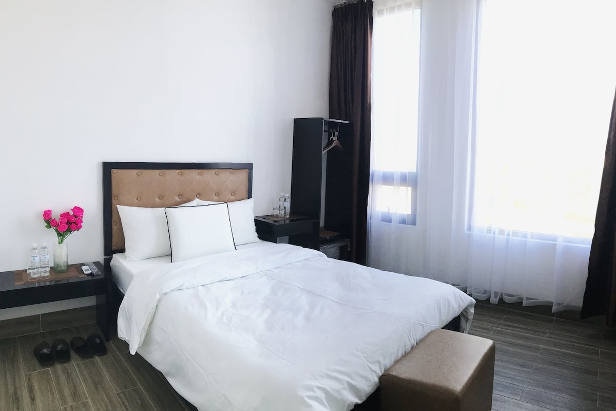 Habitación de hotel con cama, mesita de noche con un florero, televisor y ventana con cortinas.