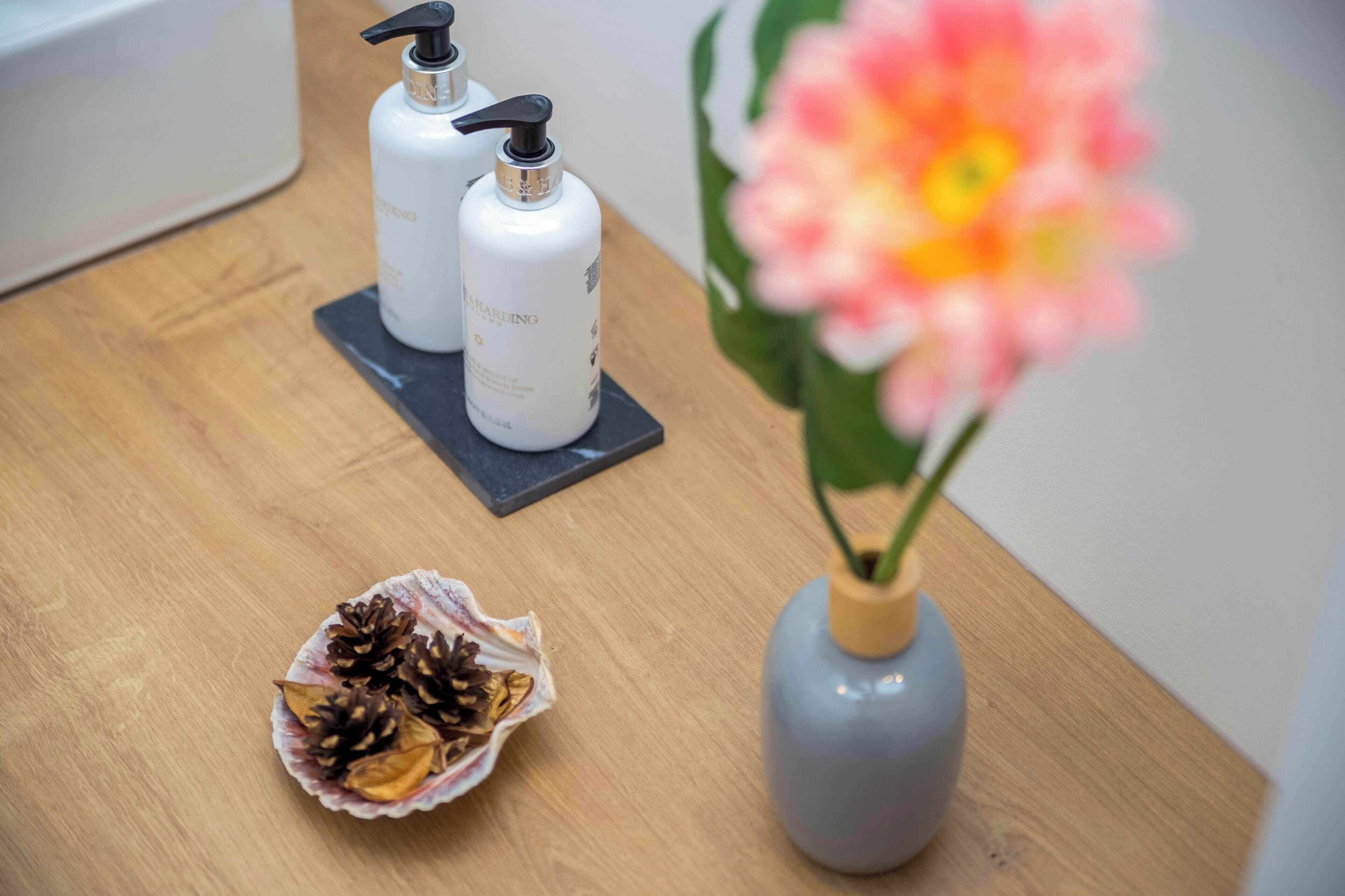 A wooden table with two white bottles, a pine cone in a paper cup, and a vase with a flower.
