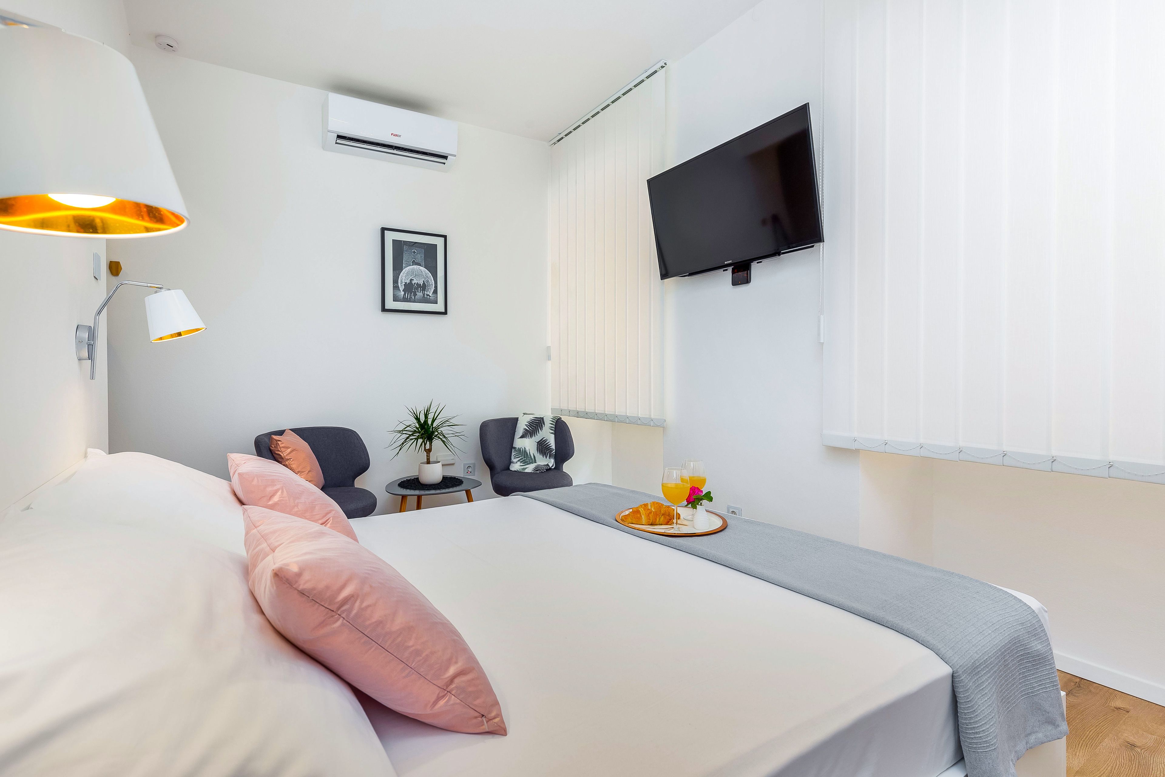 A neatly arranged hotel room with a bed, a desk, a chair, a TV, and a small table with a tray of breakfast.