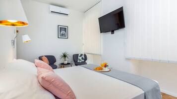 A neatly arranged hotel room with a bed, a desk, a chair, a TV, and a small table with a tray of breakfast.