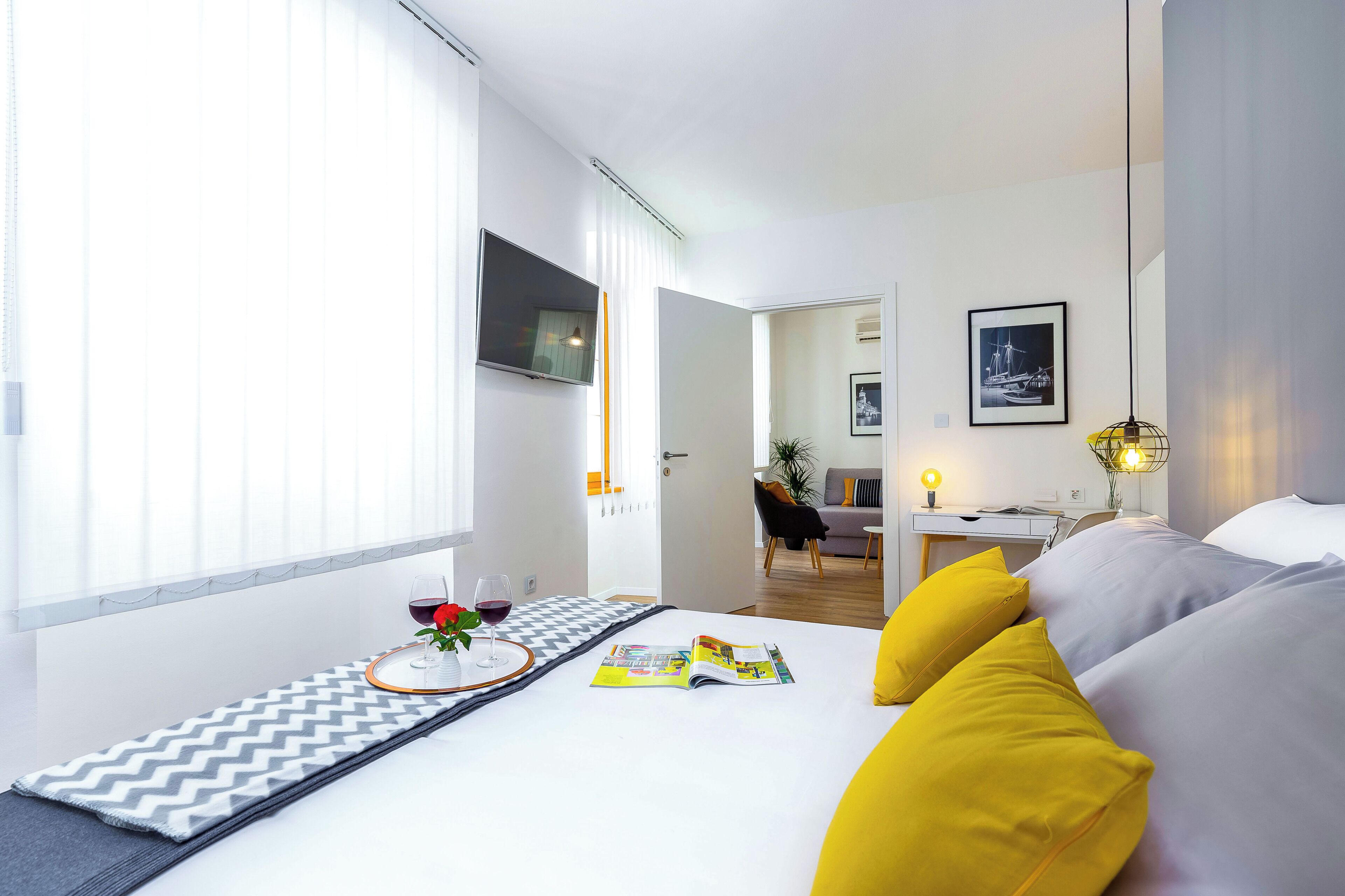 Een moderne hotelkamer met een bed, een zithoek met een tijdschrift, een klein tafeltje met een glas wijn en uitzicht op de aangrenzende woonkamer.