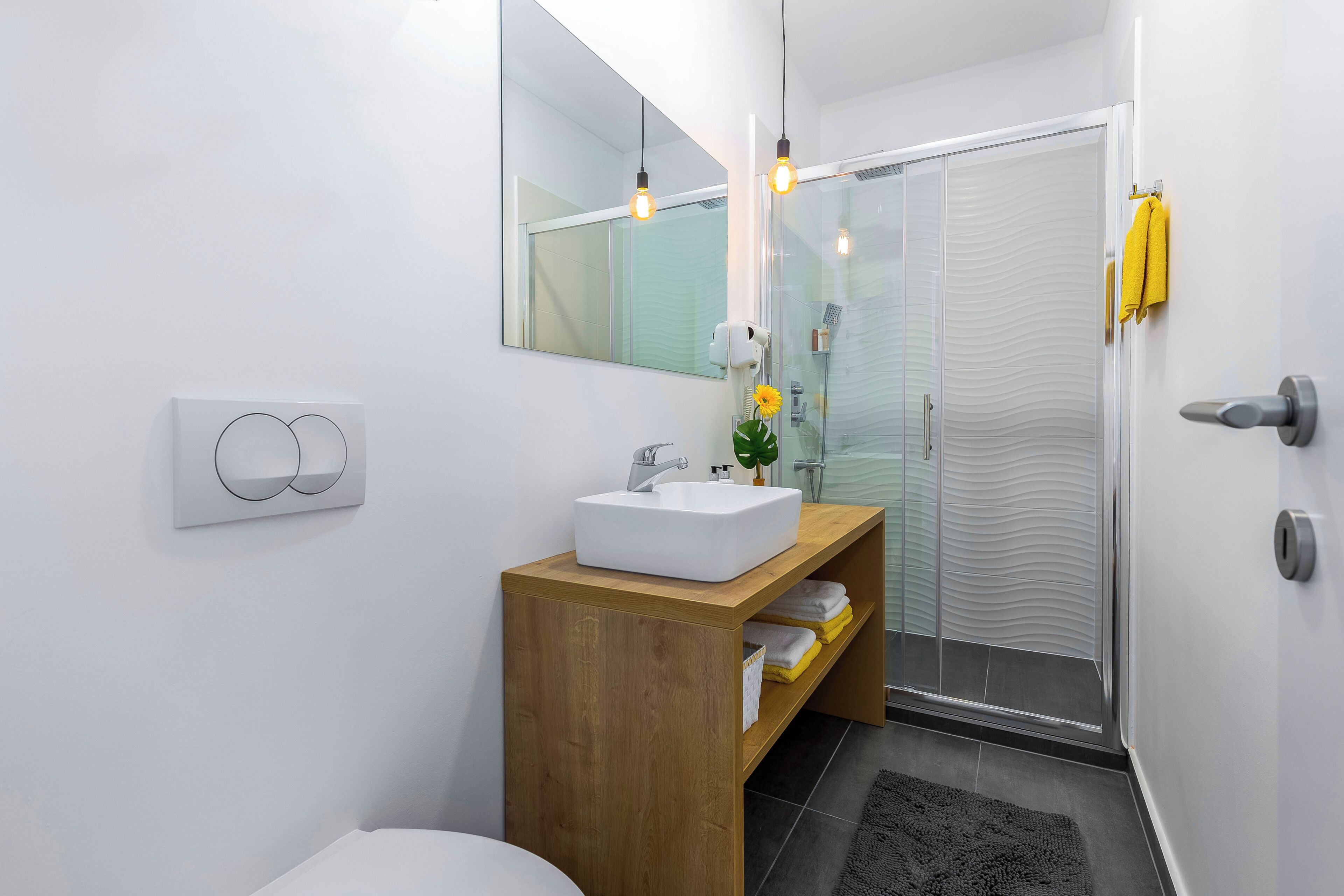 A modern bathroom with a white sink, wooden vanity, a glass shower enclosure, and a towel rack with yellow towels.