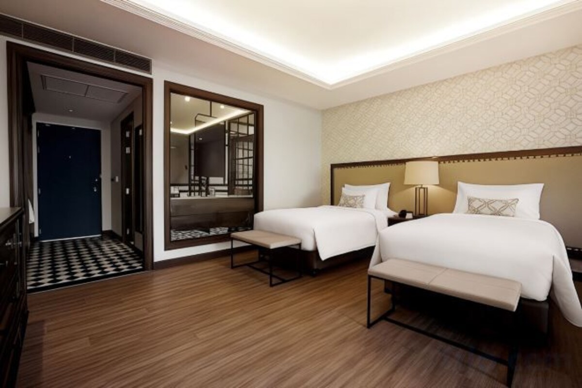 A hotel room with two beds, a large mirror, a bench, and a wooden floor.