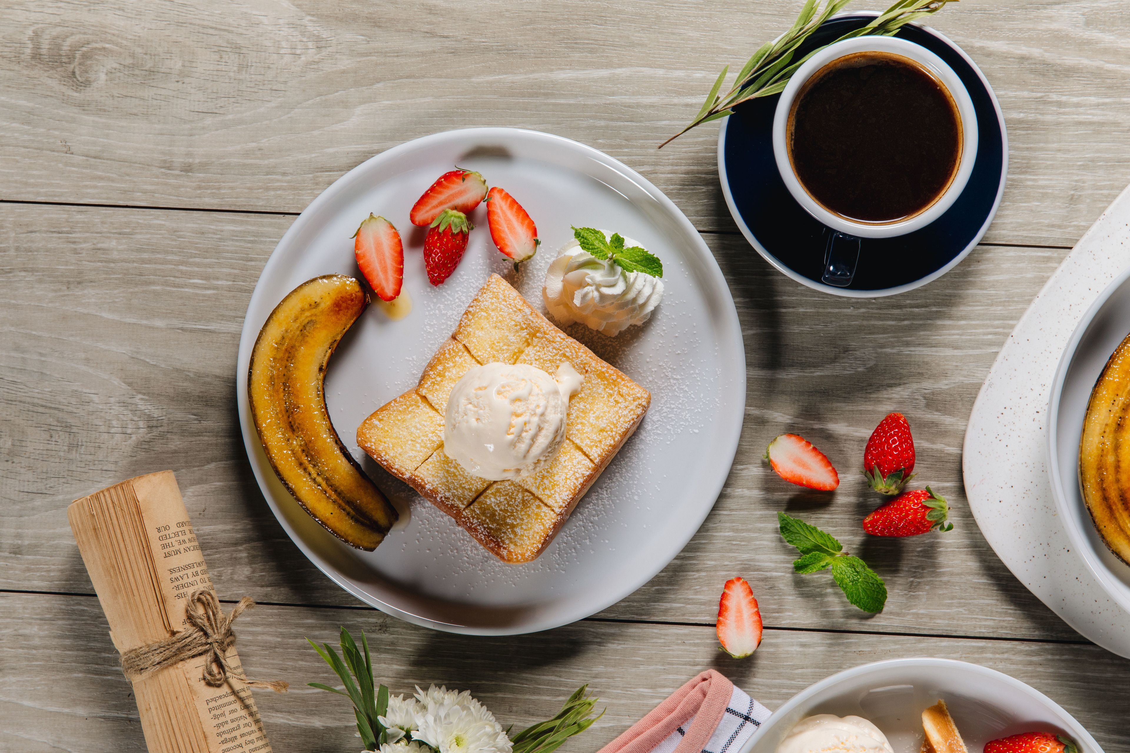 Et morgenbord med ristet brød, en banan, jordbær og flødeskum, en kop kaffe og en lille skål is.