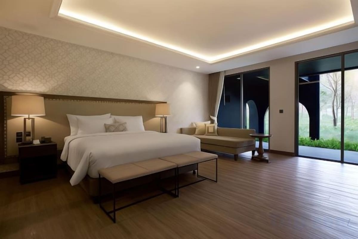 A modern hotel room with a large bed, bedside lamps, a bench, and a view through large windows.