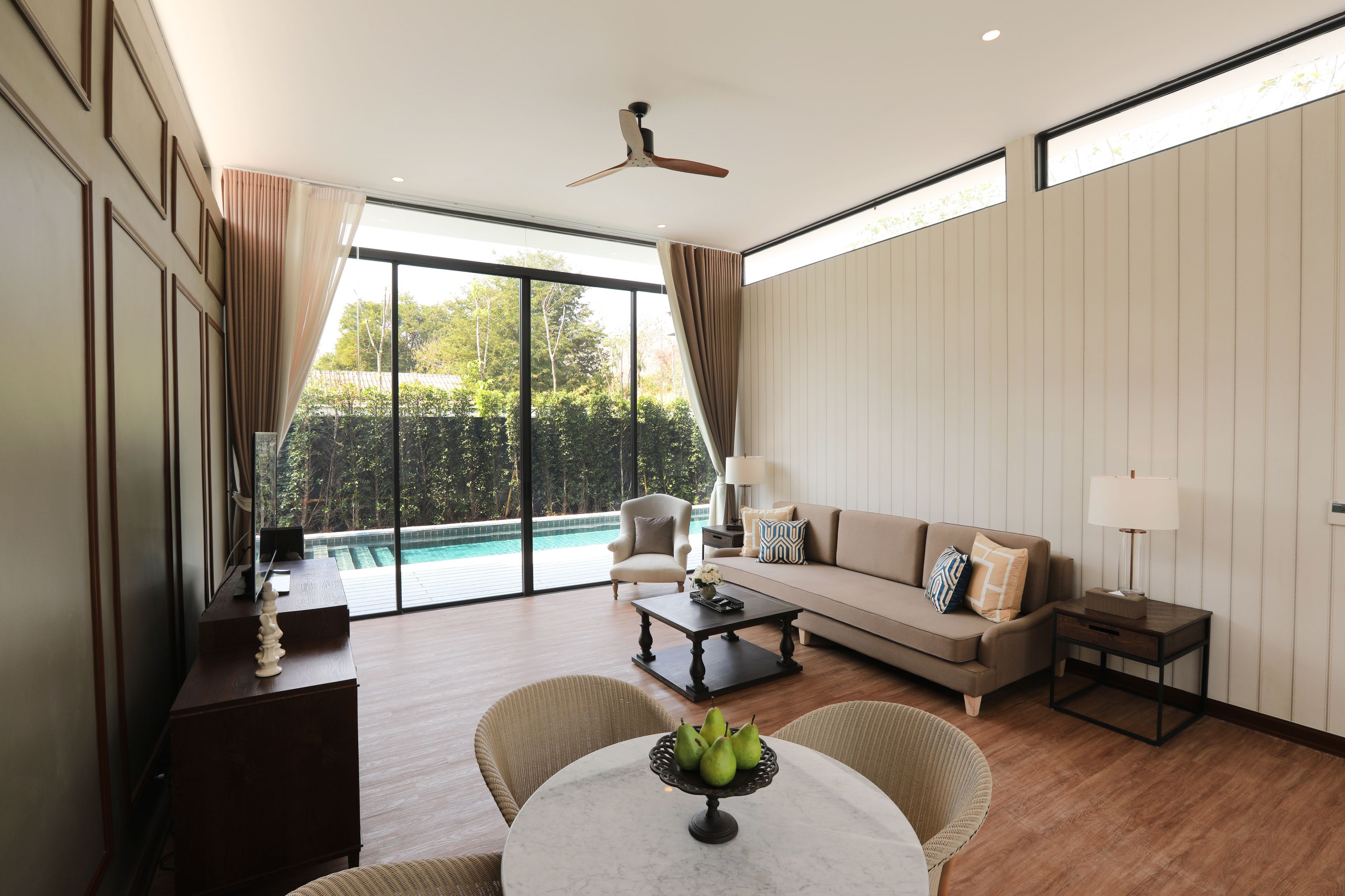 A spacious living room with a large window, a ceiling fan, a sofa, a coffee table, and a dining table with a fruit centerpiece.