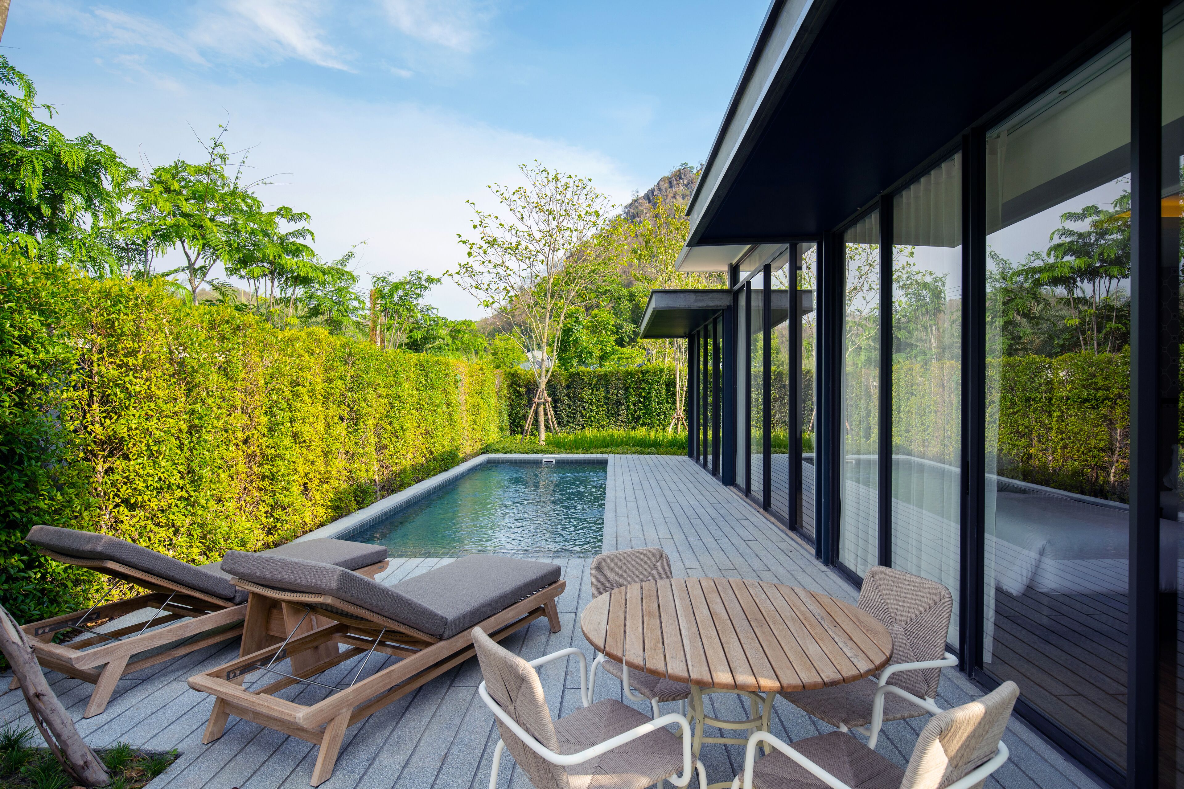 An outdoor pool area with lounge chairs and a table, surrounded by a wooden deck and glass walls.