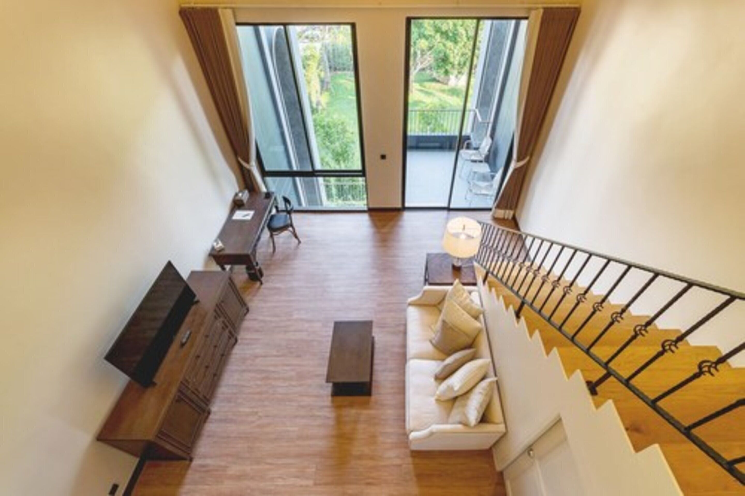 A spacious living area with a wooden floor, a sofa, a coffee table, and a desk with chairs. There is a staircase with a railing leading to an outdoor balcony.
