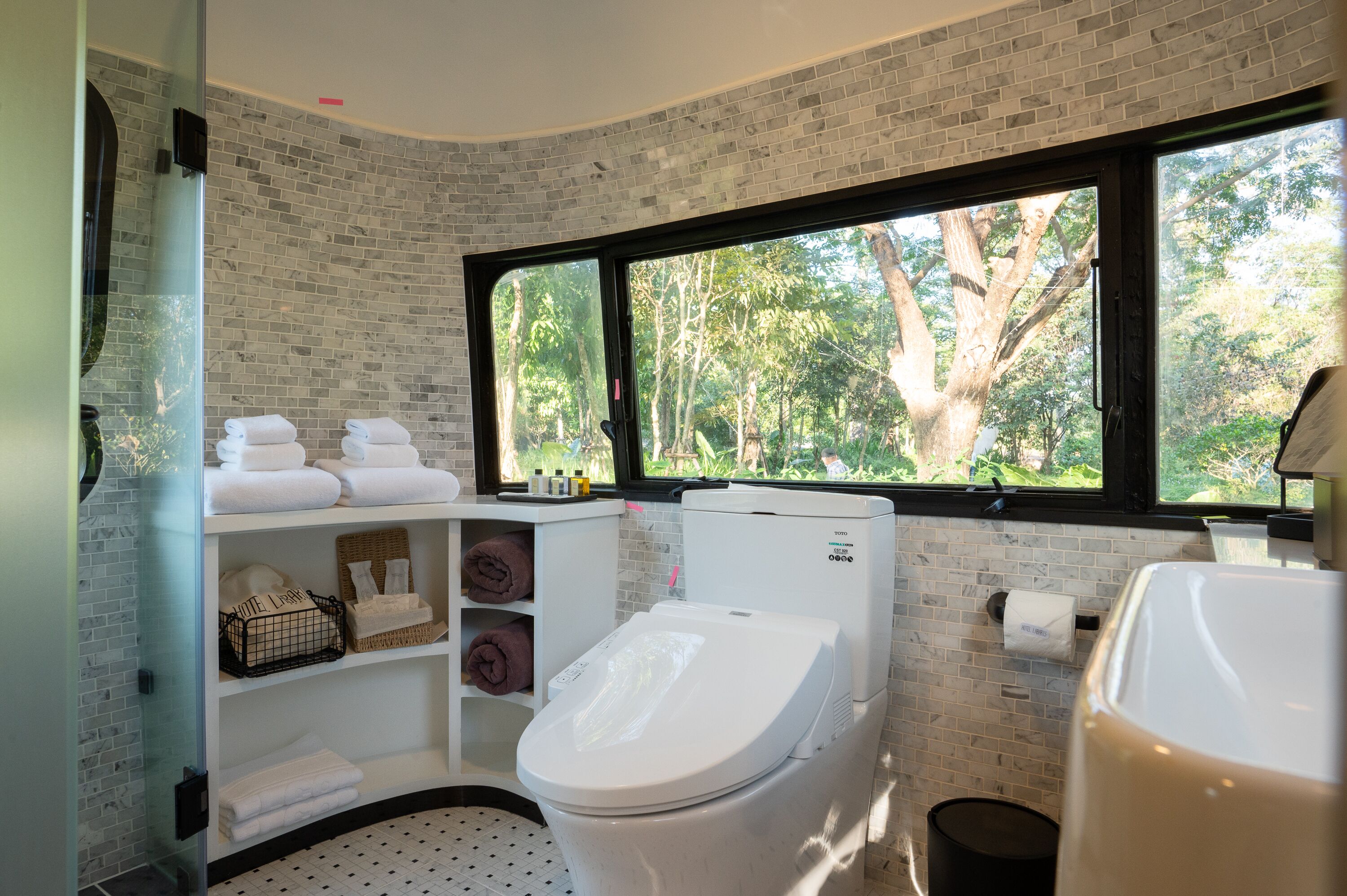 A modern bathroom with a white toilet, a glass shower enclosure, and a large window offering a view of trees.