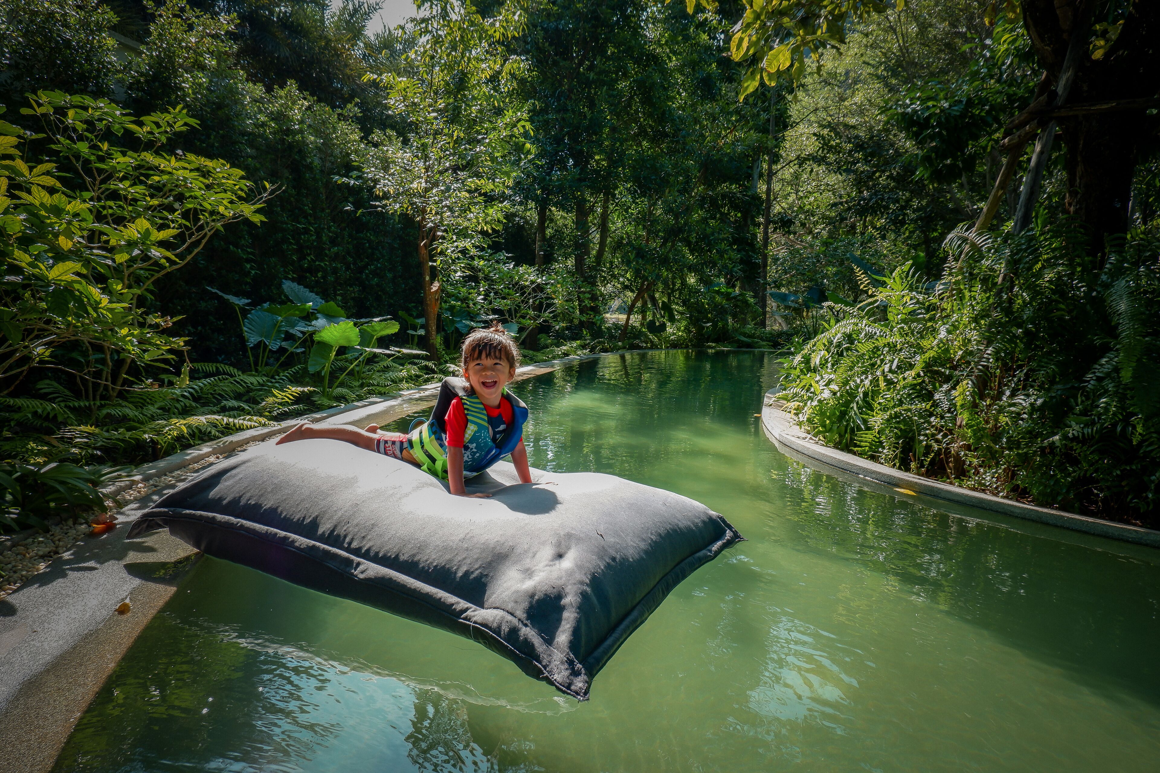 Una persona sentada sobre un cojín inflable negro en un cuerpo de agua rodeado de vegetación densa.