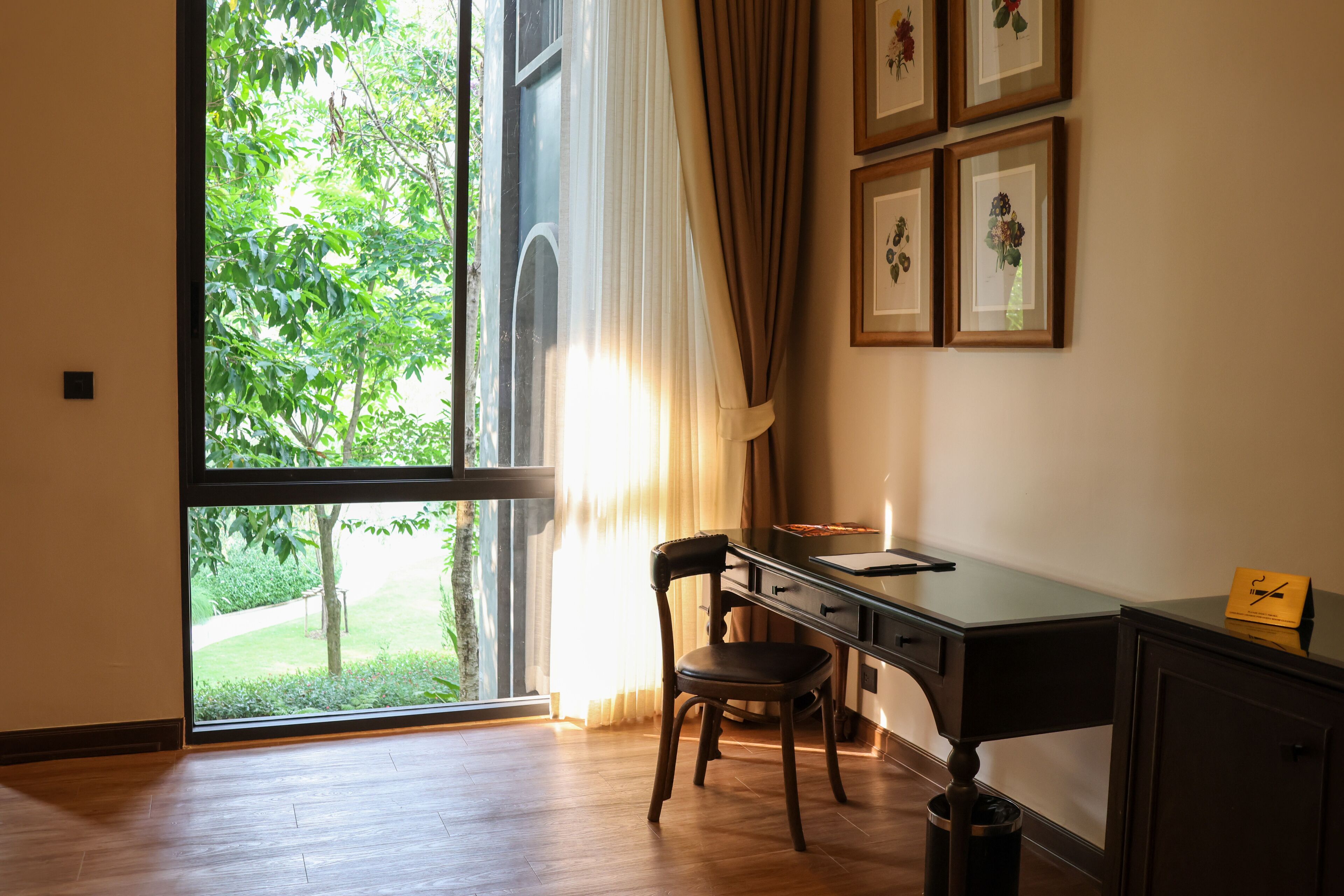 A room with a large window, a desk, a chair, and framed pictures on the wall.