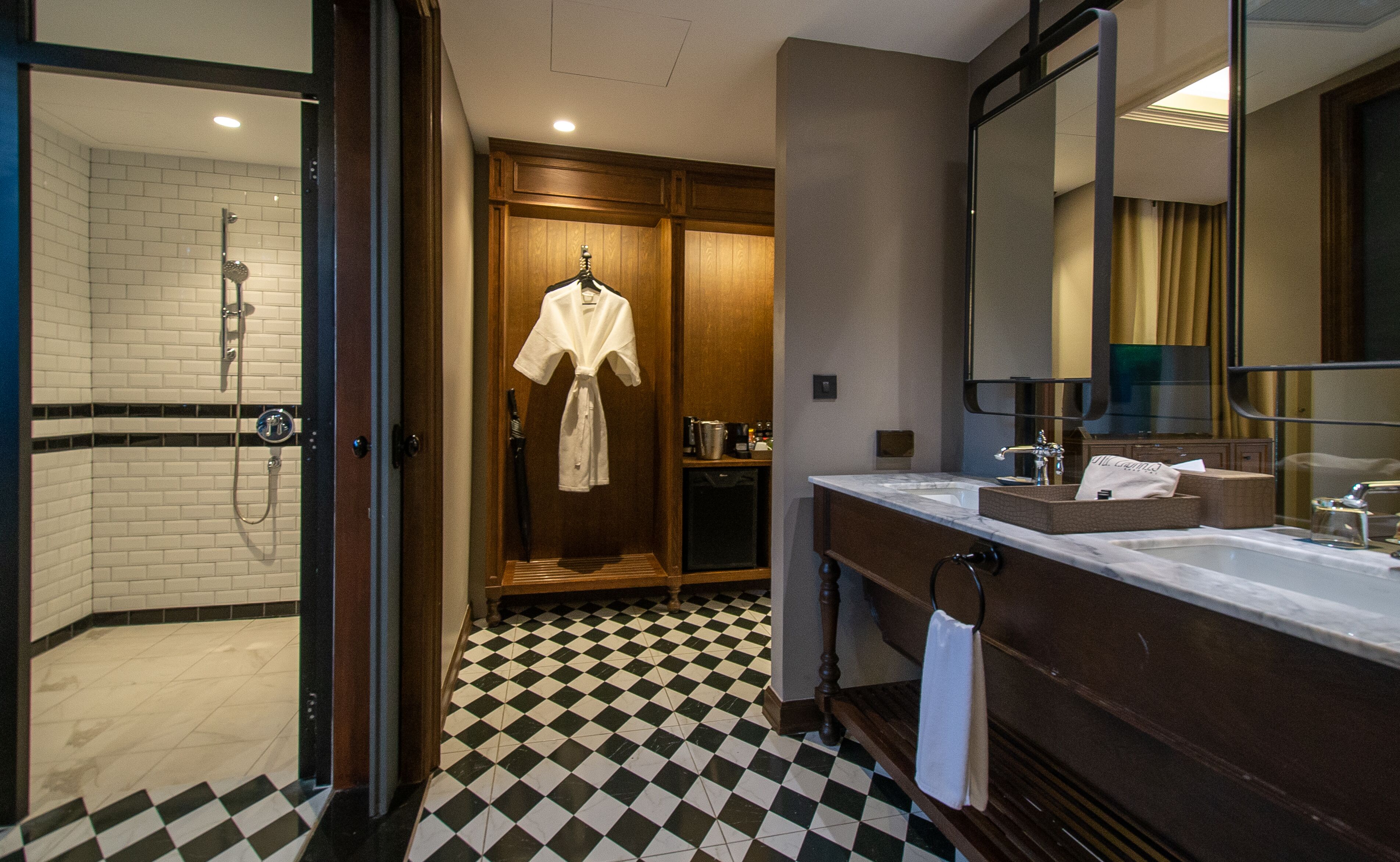 A hotel bathroom with a checkered floor, a shower, a robe hanging, a sink, and a mirror.
