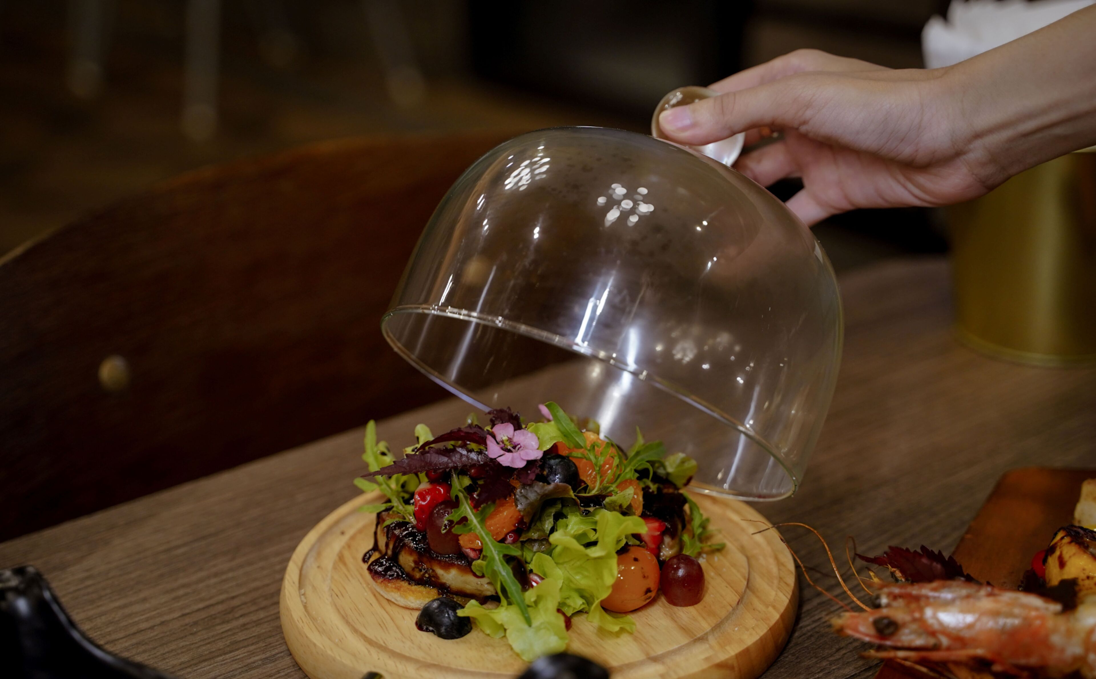 A person pouring a liquid from a glass container onto a wooden board with a salad.