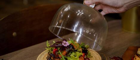 A person pouring a liquid from a glass container onto a wooden board with a salad.